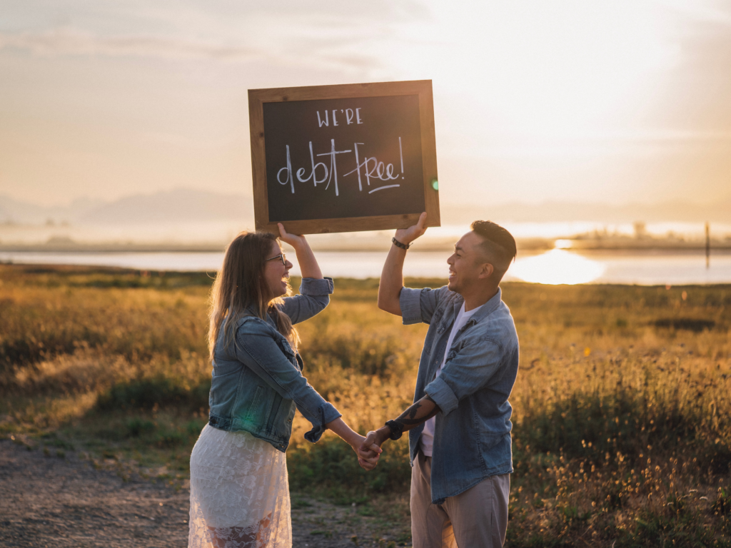 A married couple holding a sign that say's, "we're debt free!" at sunset
