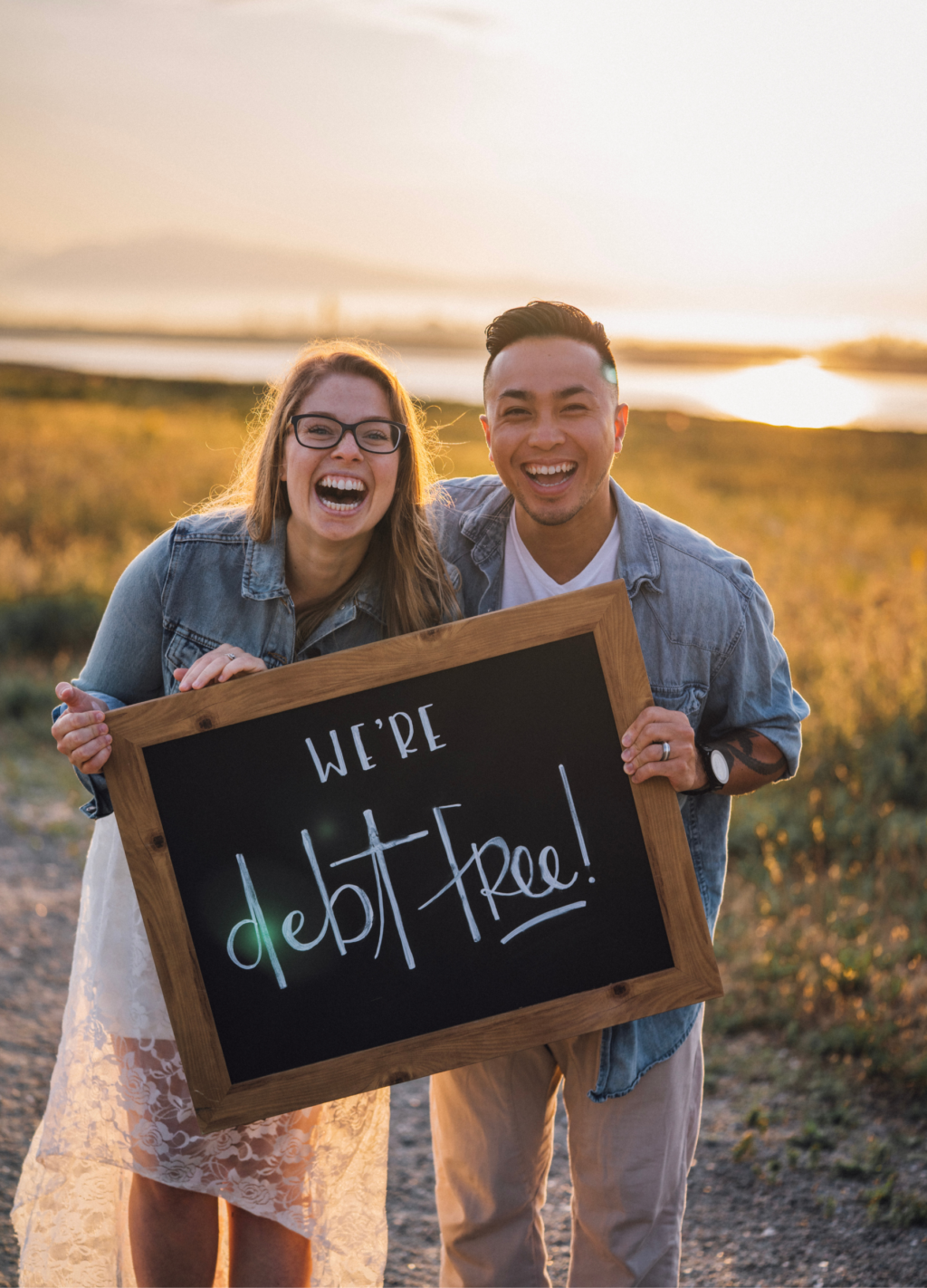A married couple holding a sign that say's, "we're debt free!" at sunset